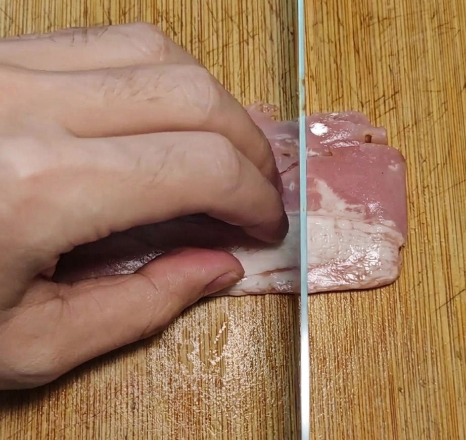Bacon strips being cut into uniform rectangular pieces on a wooden board with a kitchen knife.