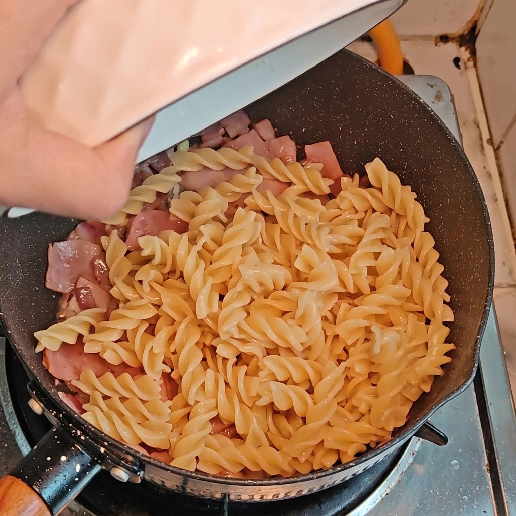 Fusilli pasta and cut asparagus pieces being added to a pan with sautéed bacon and onions.