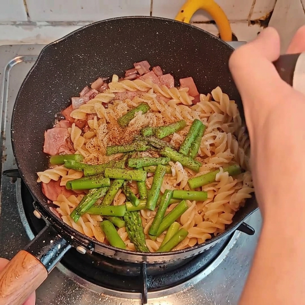 Various seasonings being sprinkled and poured over the pasta, bacon, and asparagus in the pan.