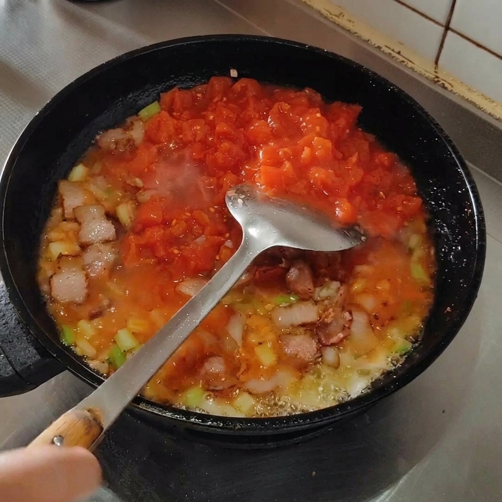 Spatula crushing tomatoes in a pan to break them down into a sauce.
