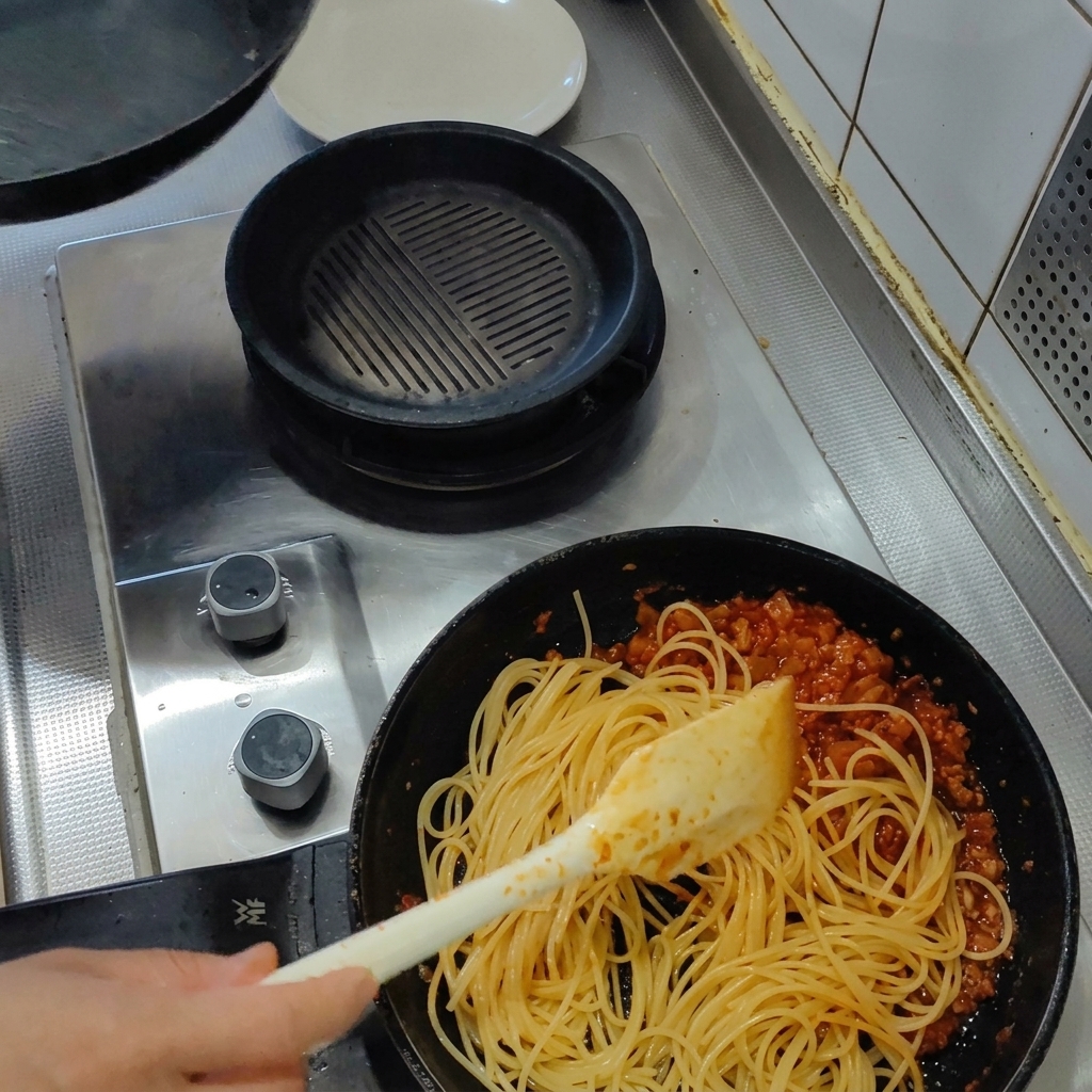 Cooked spaghetti being stirred into a pan of thick red tomato and bacon sauce with a spatula.
