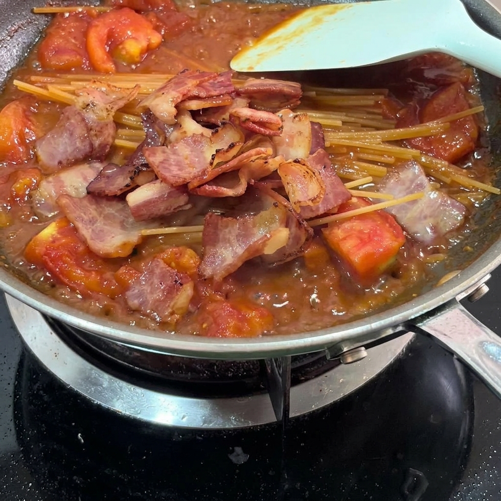 Fried bacon pieces scattered over pasta and tomato sauce in a pan.