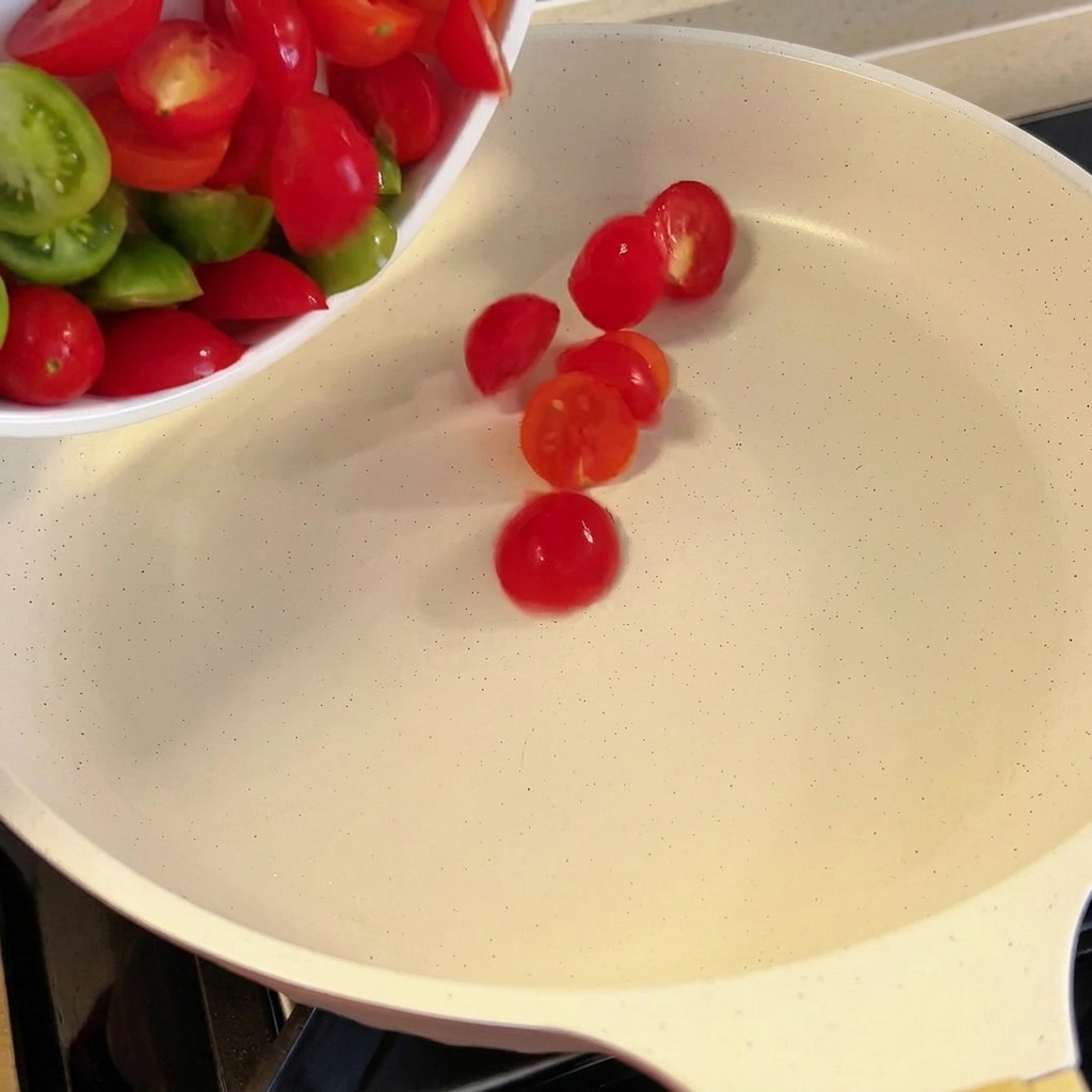 Fresh halved cherry tomatoes being added to a white skillet.