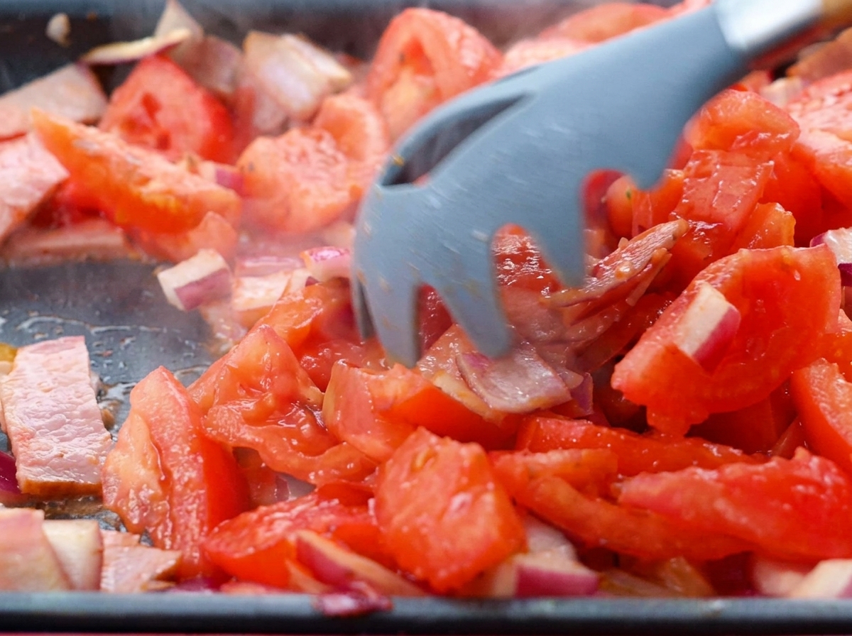 Diced tomatoes being stirred into a bacon and onion mixture in a pan.