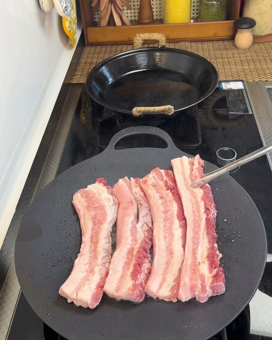 Tongs flipping strips of pork belly in a pan, revealing the cooked, browned side.