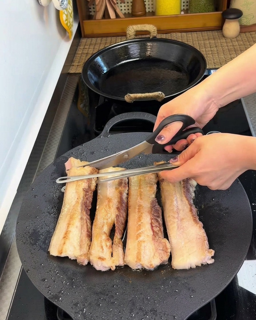 Kitchen scissors cutting browned pork belly strips into smaller chunks inside the cast iron pan.