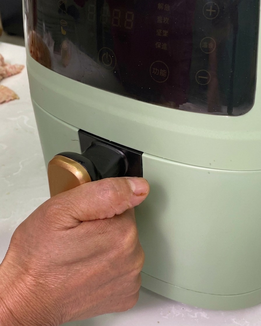 A hand sliding an aluminum pan filled with seasoned pork rolls into a green air fryer.