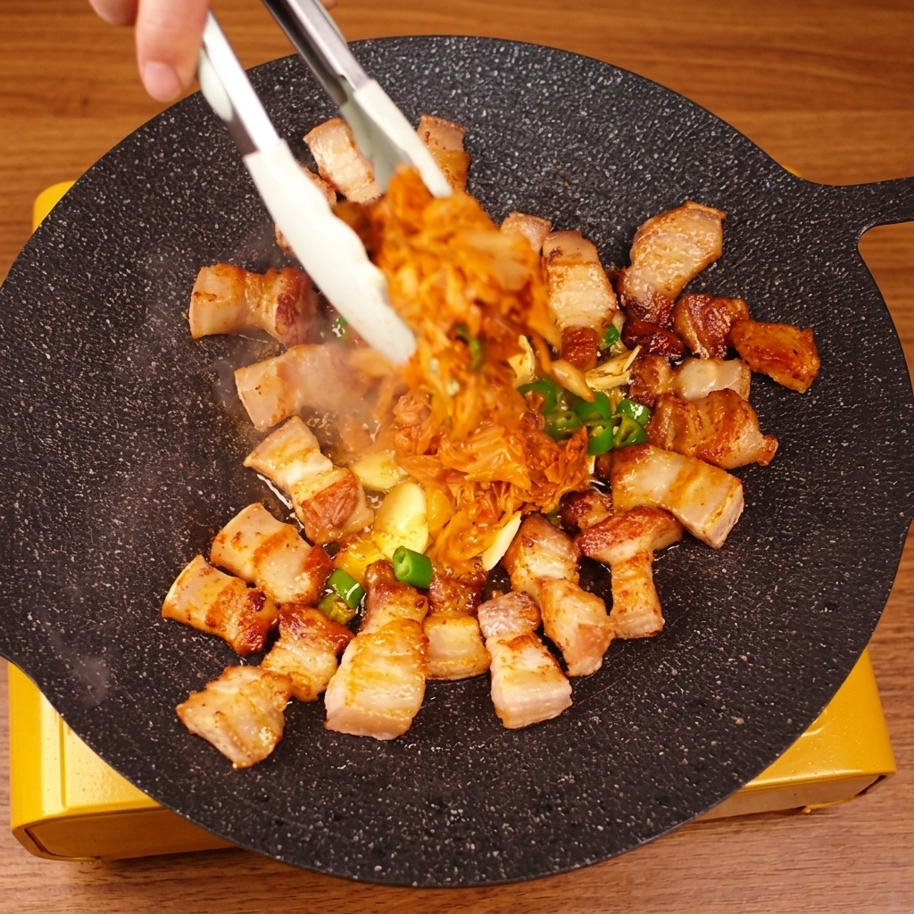 Hand using tongs to toss pork belly and kimchi together on a hot griddle.