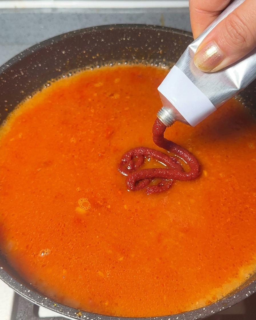 Tomato paste being squeezed from a metal tube into a simmering orange-red soup base.