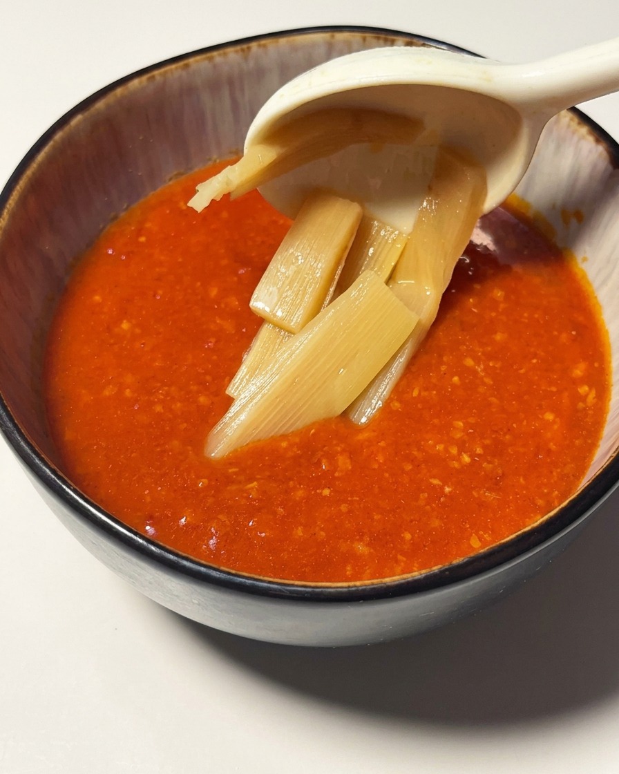 Slices of bamboo shoots being lifted by a spoon into a bowl filled with vibrant tomato shrimp sauce.