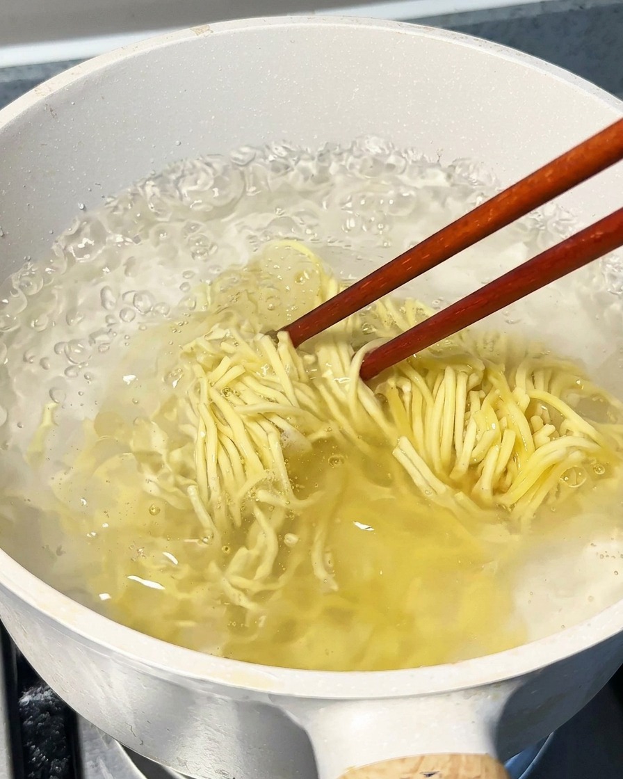 Noodles boiling in a large pot of water, being stirred with wooden chopsticks.