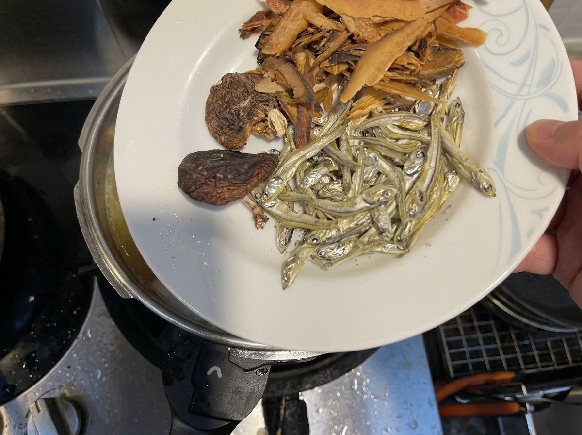 Dried fish, shiitake mushrooms, and fish flakes being dropped into a pot of simmering liquid.