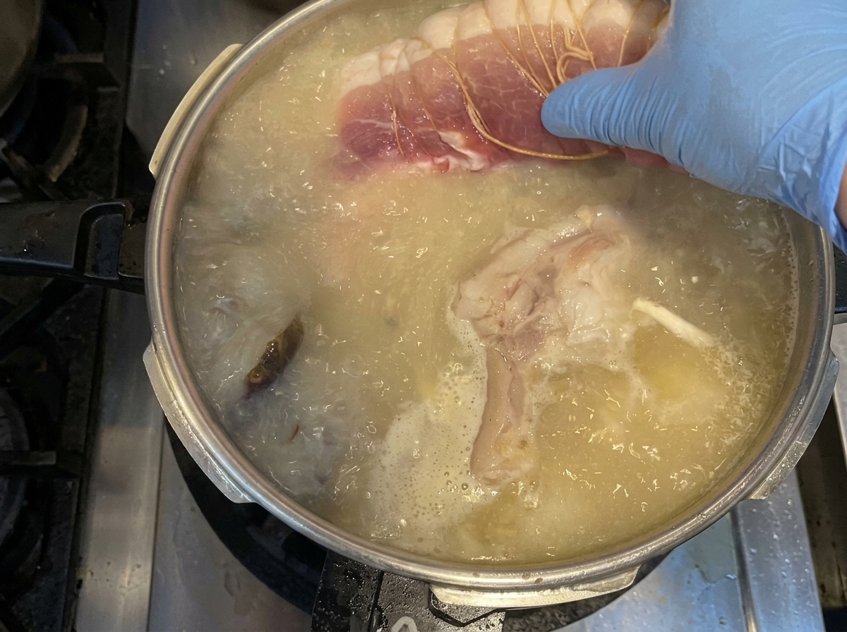 A hand wearing a blue glove placing a piece of raw, tied pork shoulder into a pot of boiling broth with vegetables.