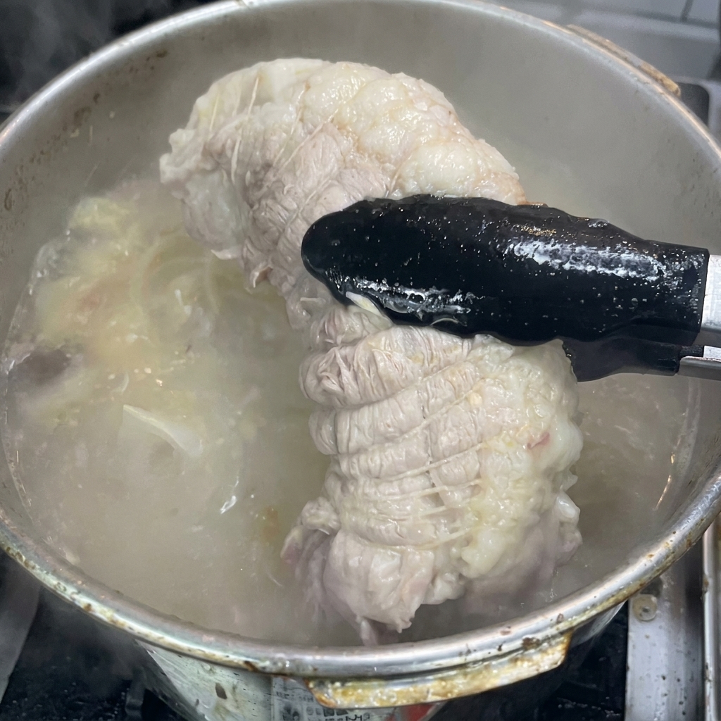 Black tongs lifting a fully cooked, tied piece of pork shoulder out of a bubbling pot of broth.