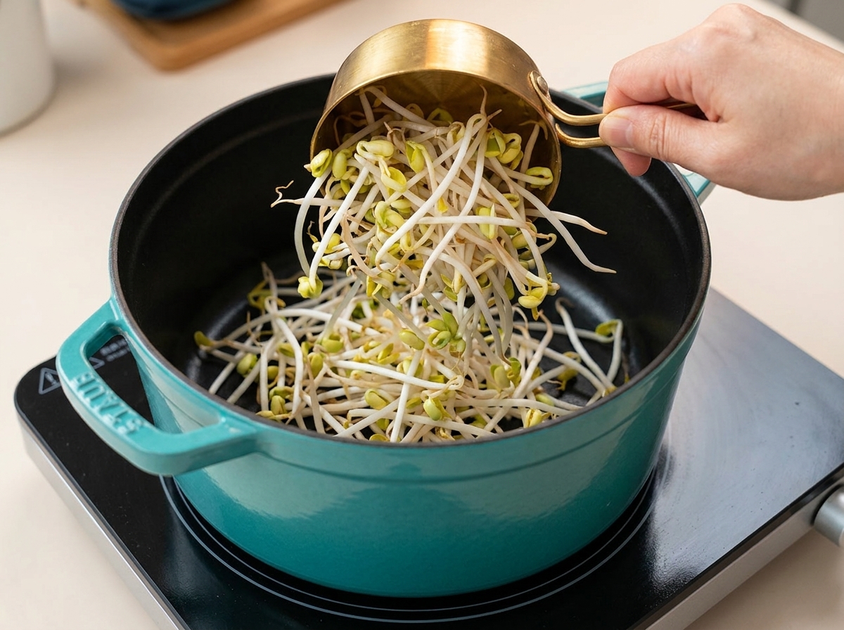 A hand pouring fresh, raw bean sprouts from a small golden measuring cup into a large teal cooking pot.