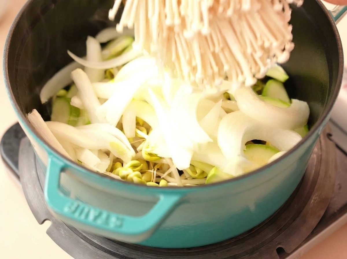 A large cluster of raw enoki mushrooms being placed into a teal pot already containing sliced white onions, green zucchini, and bean sprouts.