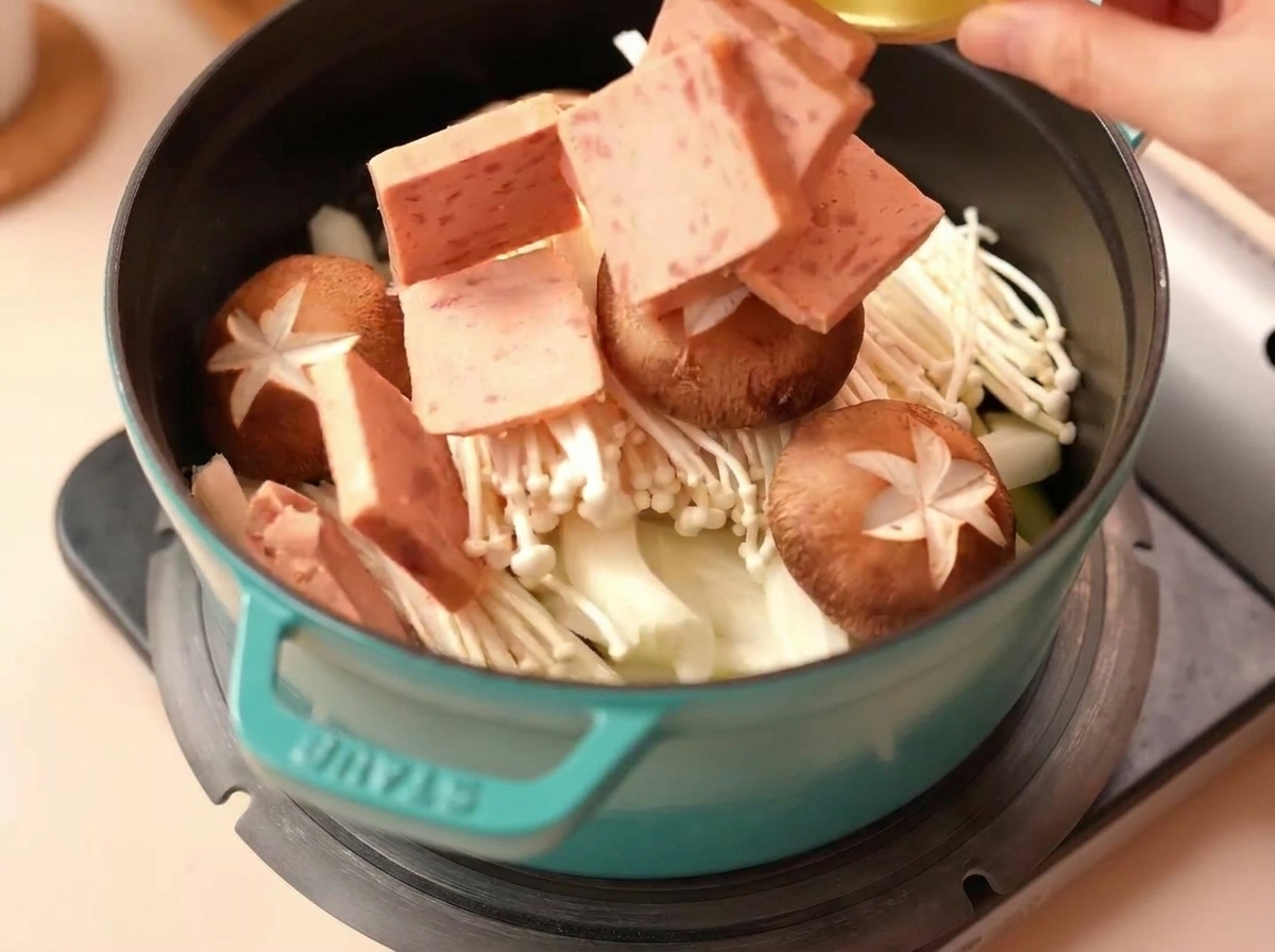Square pieces of Spam being added to a pot filled with enoki mushrooms, onions, and scored shiitake mushrooms.