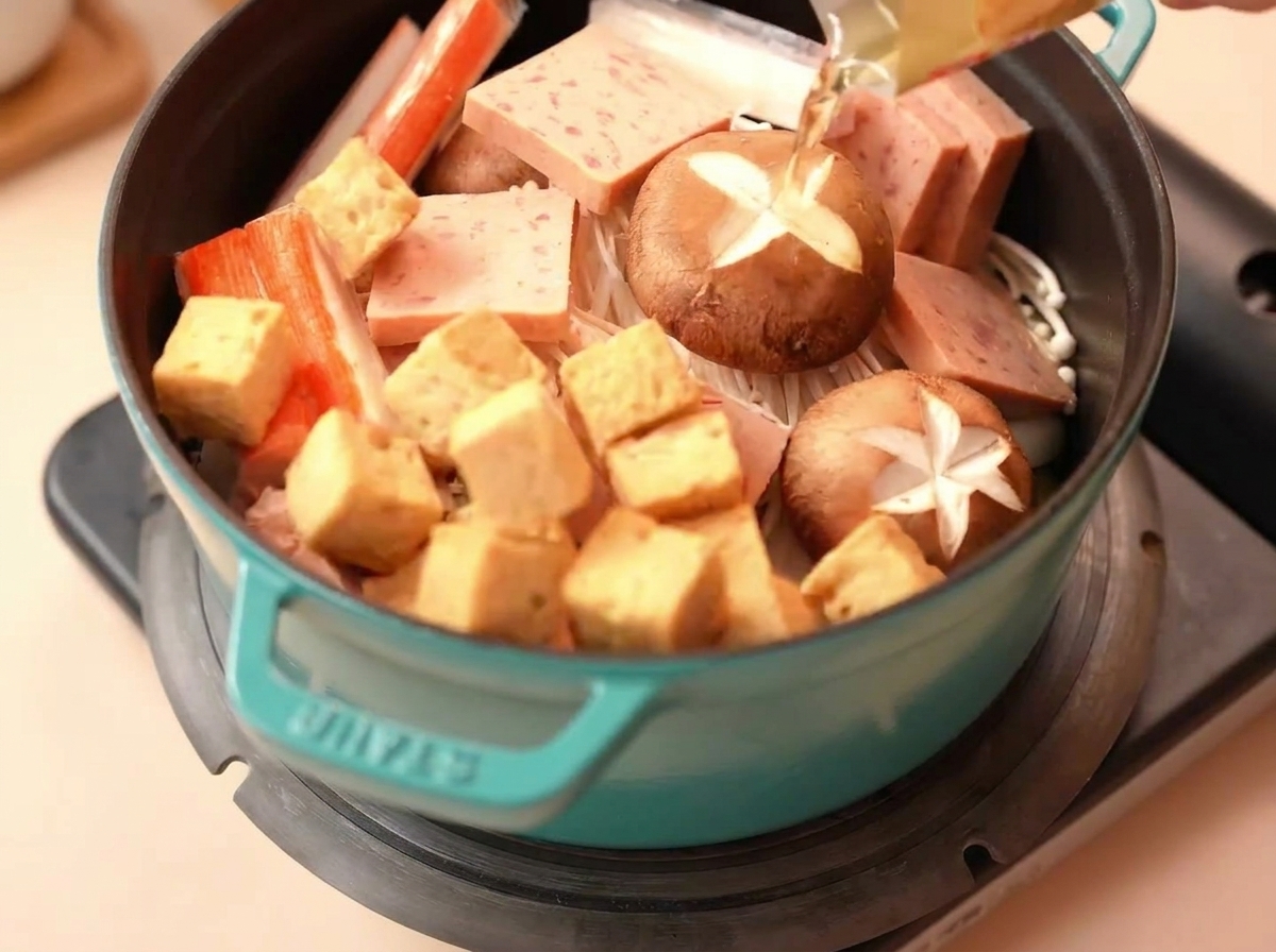 Fish tofu cubes and crab sticks placed into a pot with Spam and fresh mushrooms.