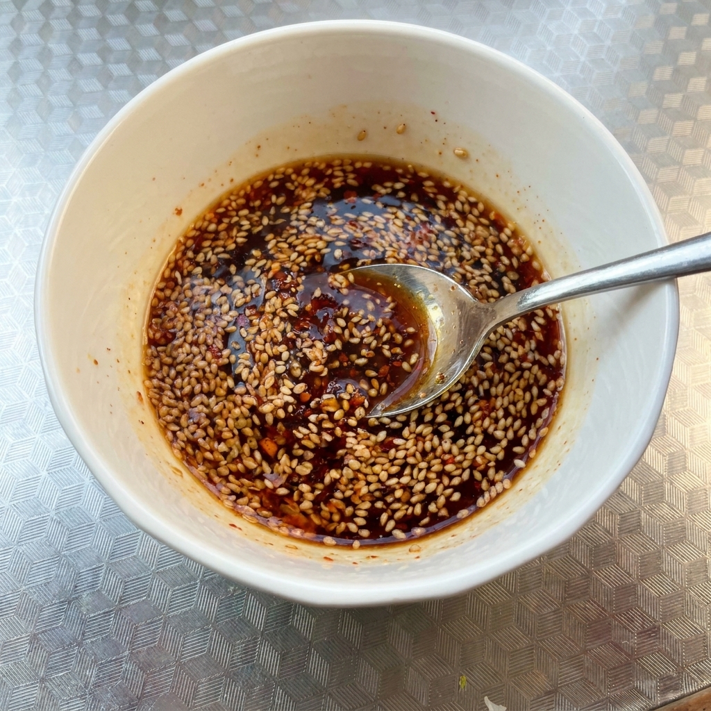 A close-up of a white bowl filled with a dark, spicy sauce mixture topped with toasted sesame seeds, being stirred with a spoon.