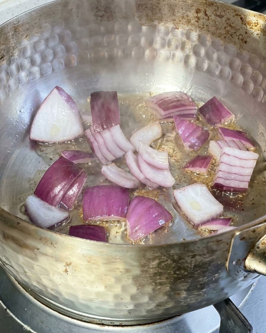 Red onion slices sizzling in a metal pan with hot oil on a stovetop.