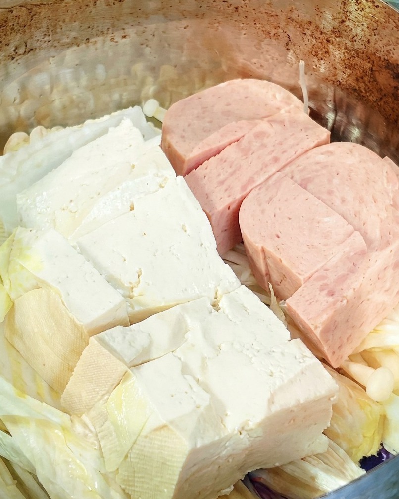 A close-up view showing a block of dried instant noodles being placed into the center of the pan on top of the other ingredients.