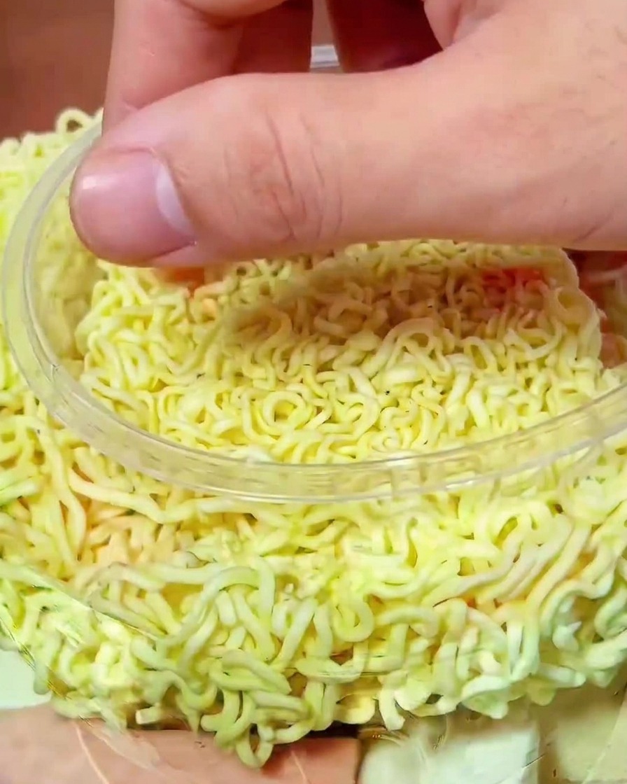 A hand placing vibrant red kimchi pieces over the uncooked noodles in the cooking pot.
