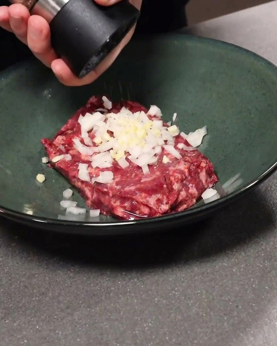 Ground beef mixed with garlic and onion in a bowl, with black pepper being added.