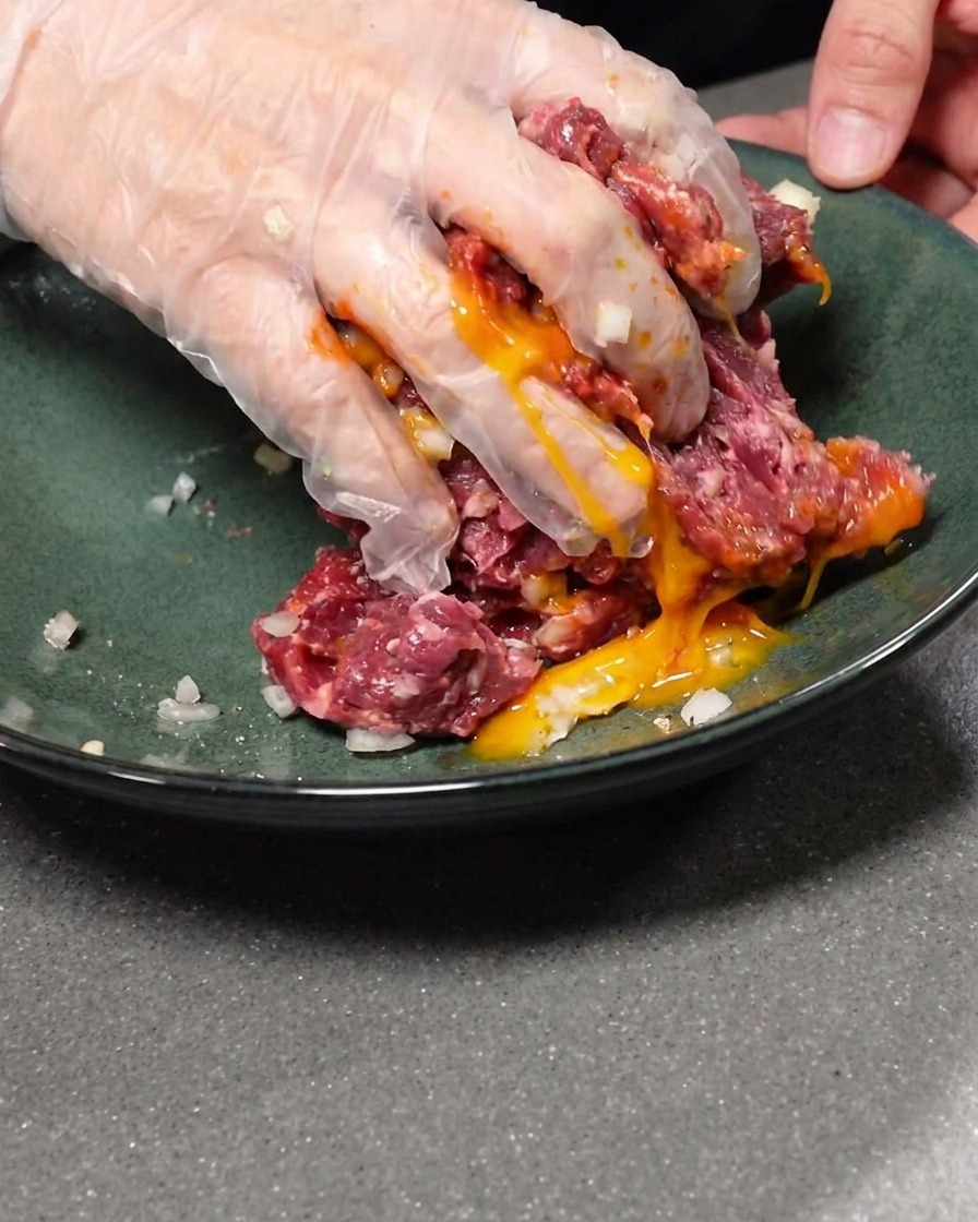 A raw egg yolk being incorporated into the seasoned ground beef mixture.