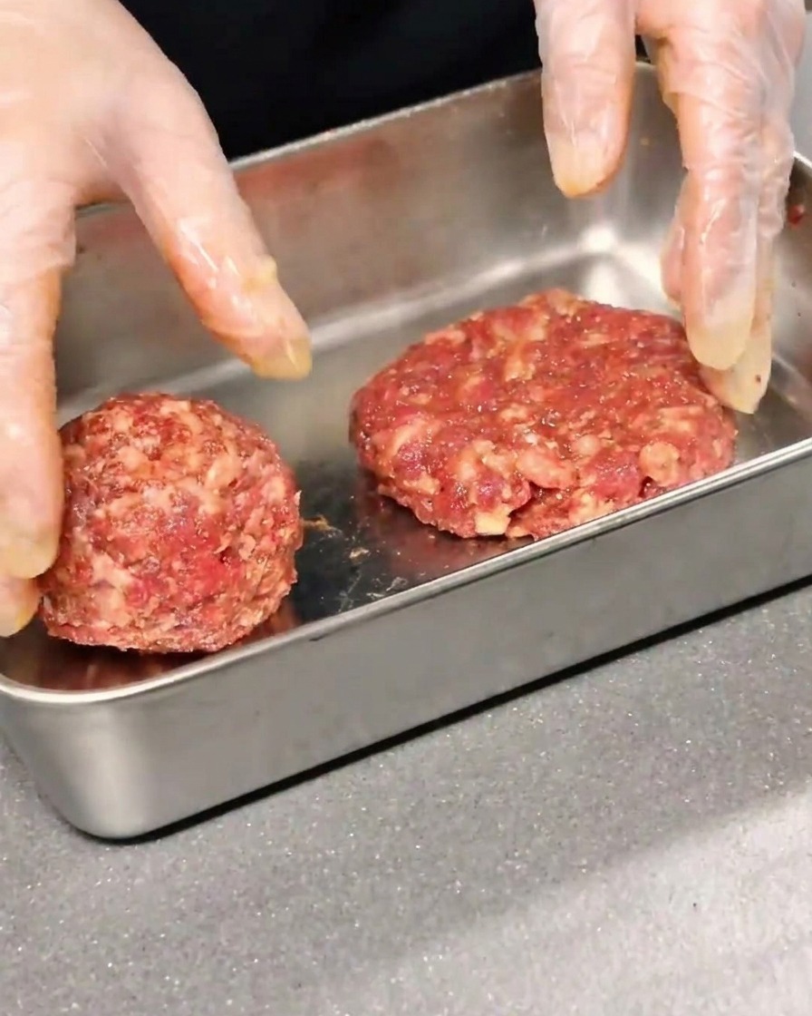 Two seasoned beef patties being shaped and flattened in a metal container.