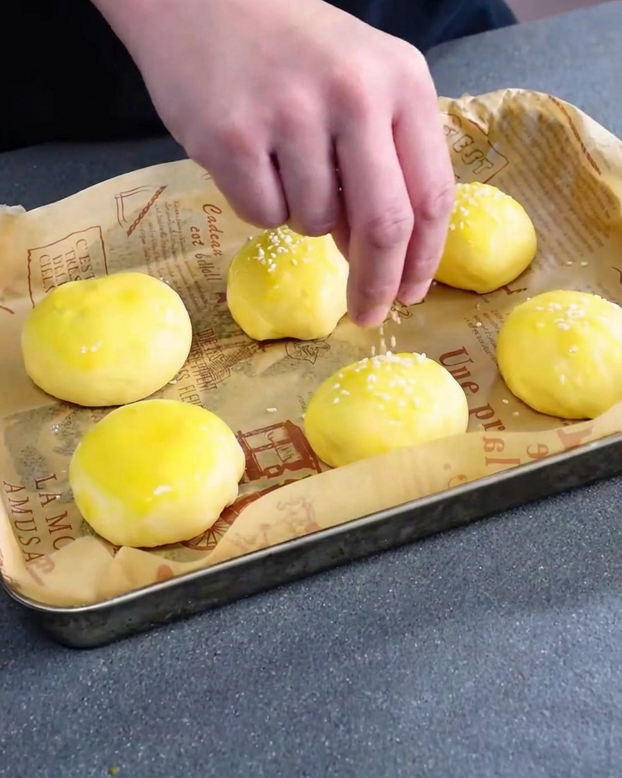White sesame seeds being sprinkled over egg-washed, unbaked burger buns.