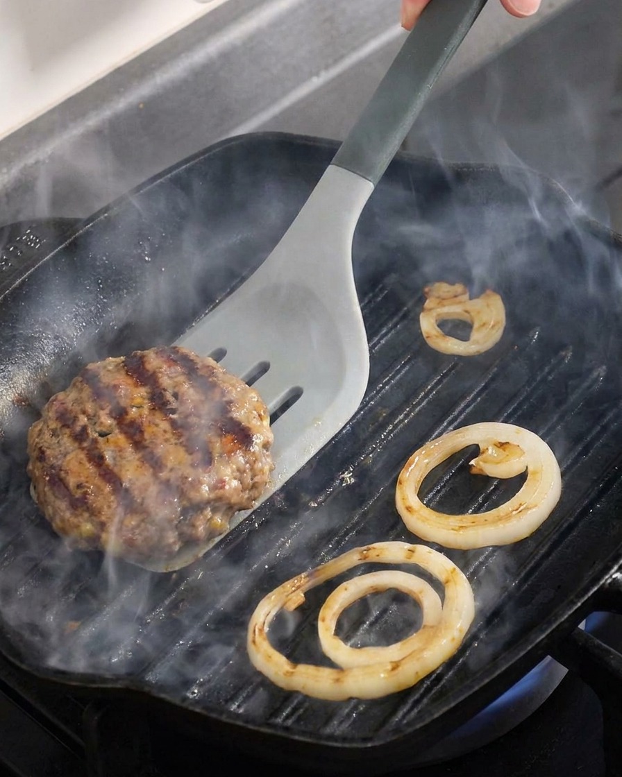 Close-up of a beef patty and onion rings being flipped on a hot grill pan.
