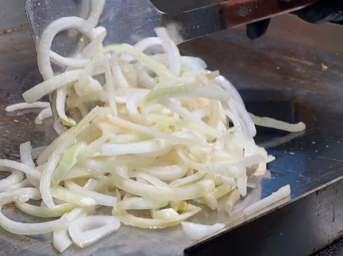 Sliced onions being spread across a hot griddle using a large metal spatula.