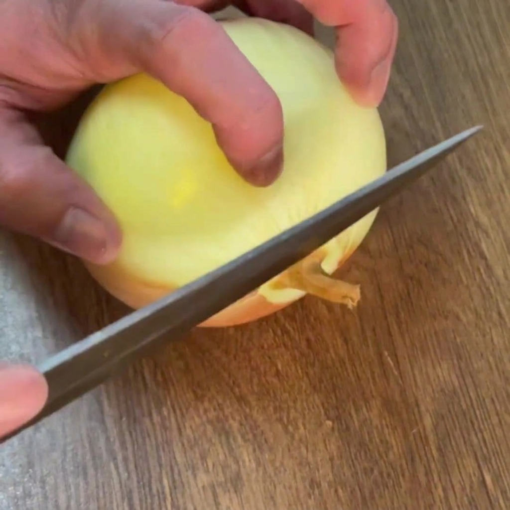 Hands using a knife to slice the root end off a peeled yellow onion on a wooden cutting board.
