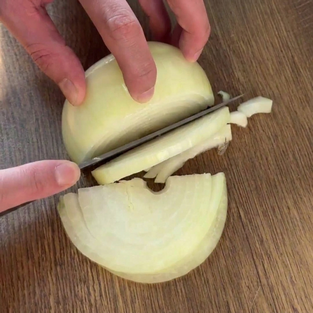 A person slicing a halved yellow onion into uniform half-ring strips with a chefs knife on a wooden board.