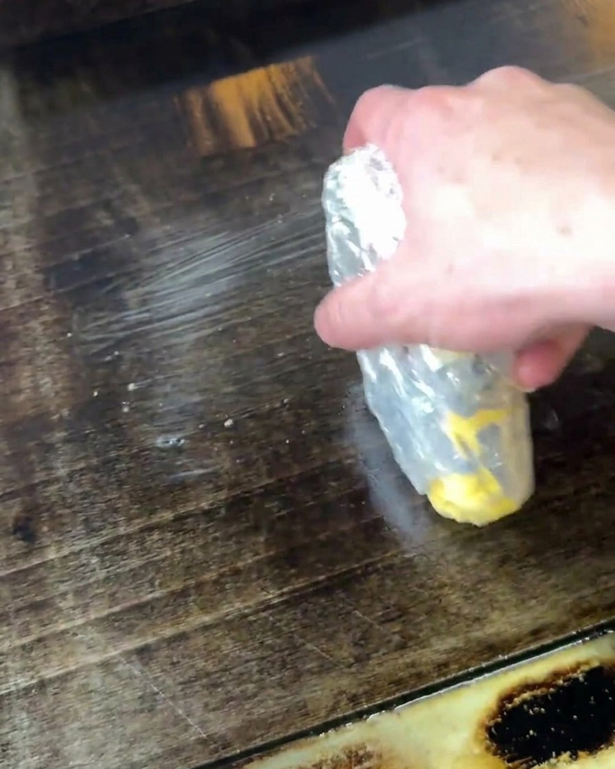 A hand holding a partially wrapped stick of butter, melting it by rubbing it directly onto a hot, dark flat-top griddle.