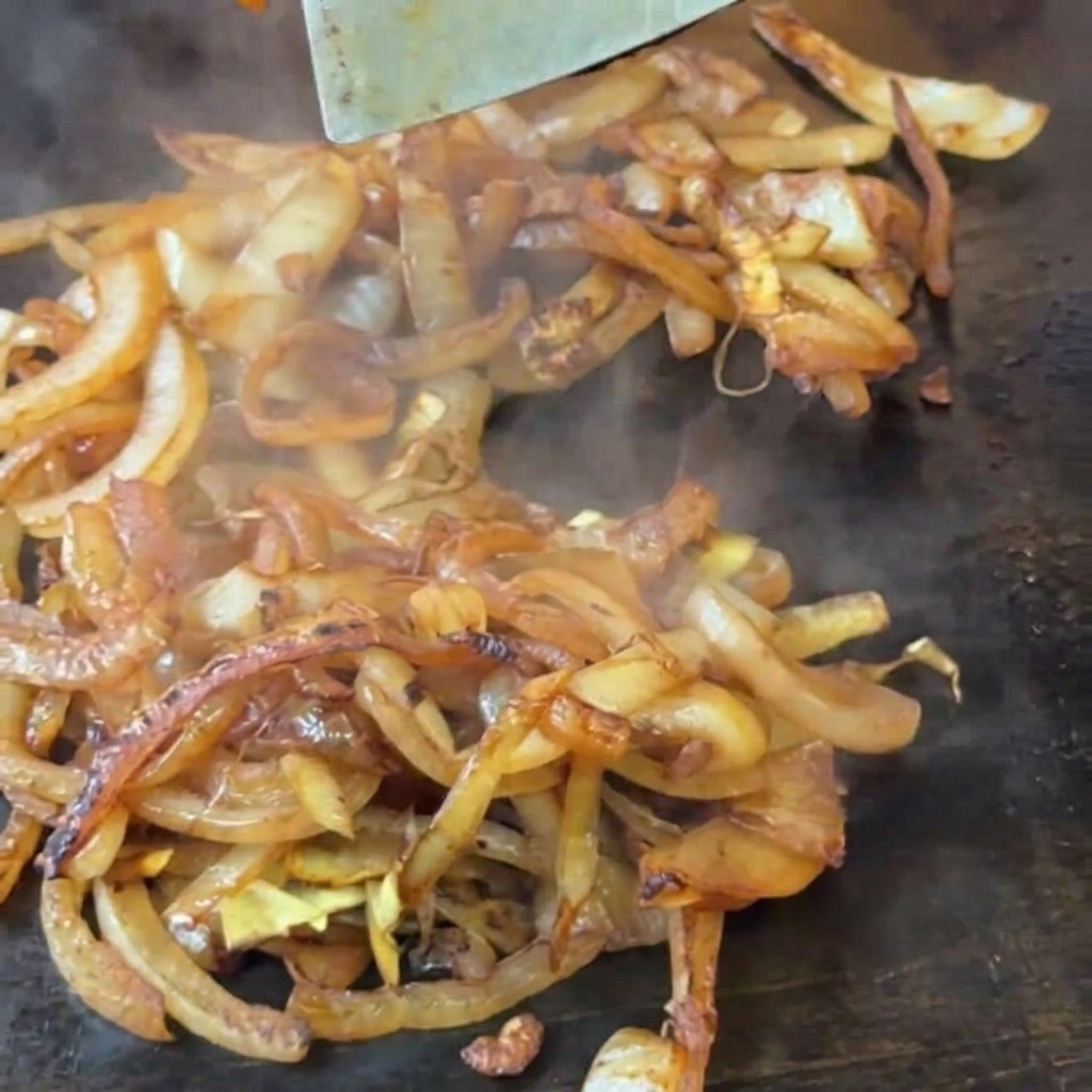 A metal spatula stirring softening, light brown sliced onions on a hot griddle.