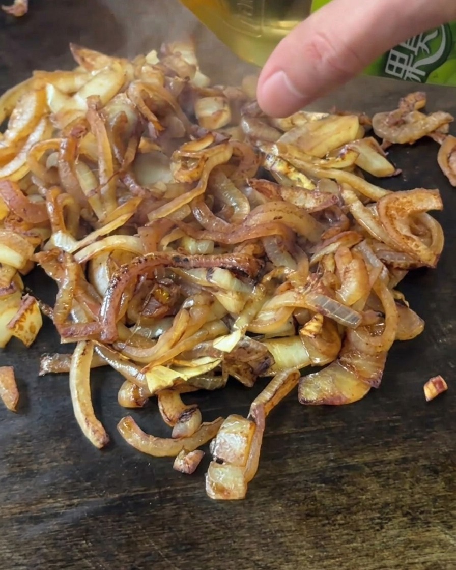 A hand pouring apple cider vinegar from a green bottle onto a pile of browned onions on a flat top.