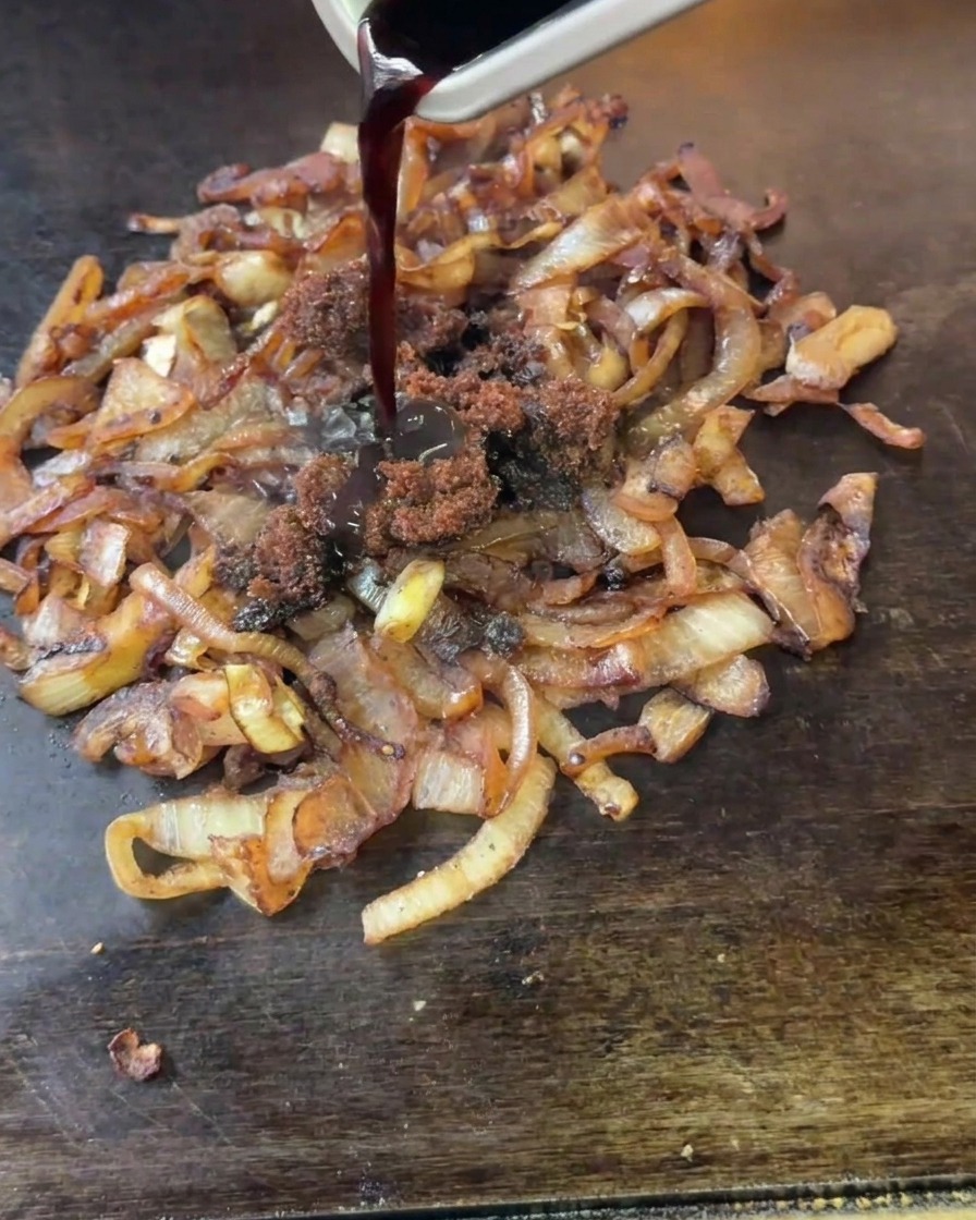 Dark mature vinegar and clumps of brown sugar being poured from a small metal container onto cooking onions.