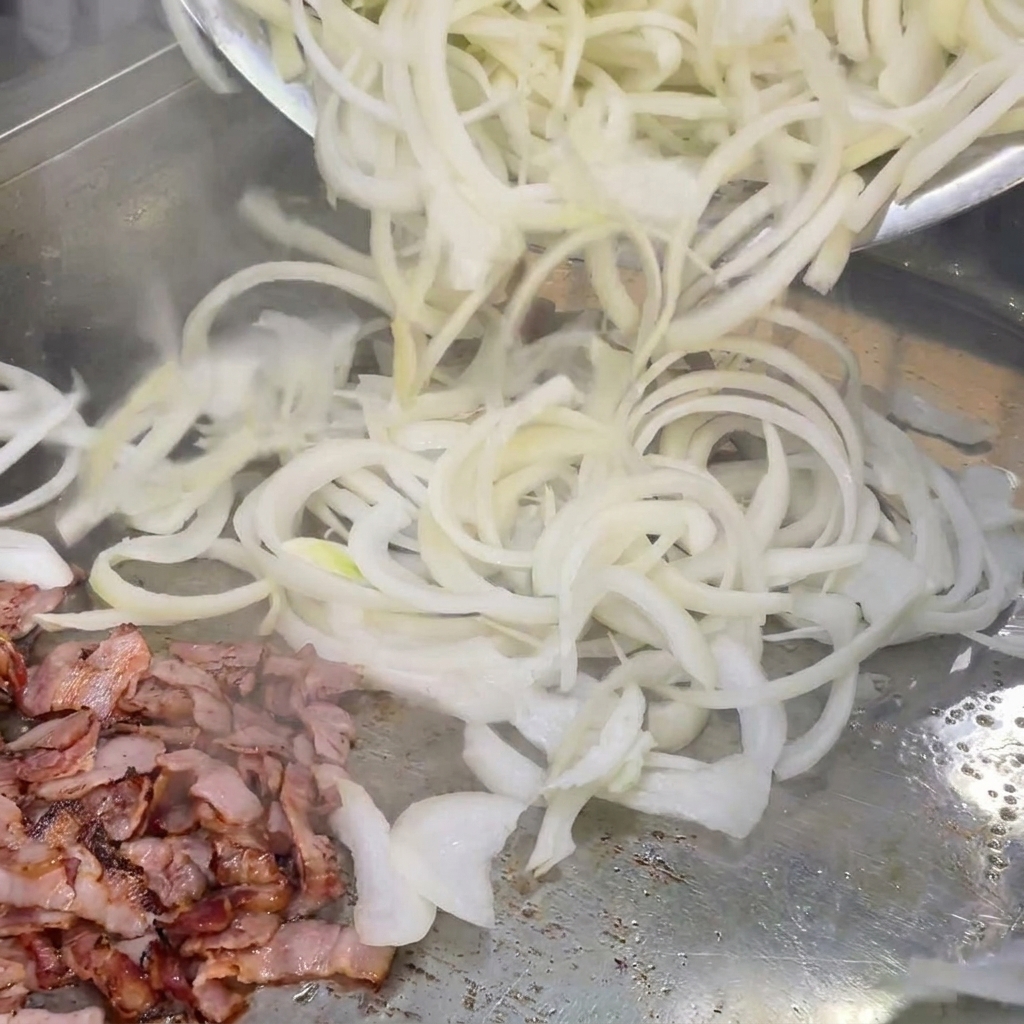 Freshly sliced white onions being tossed with cooked bacon pieces on a hot grill.
