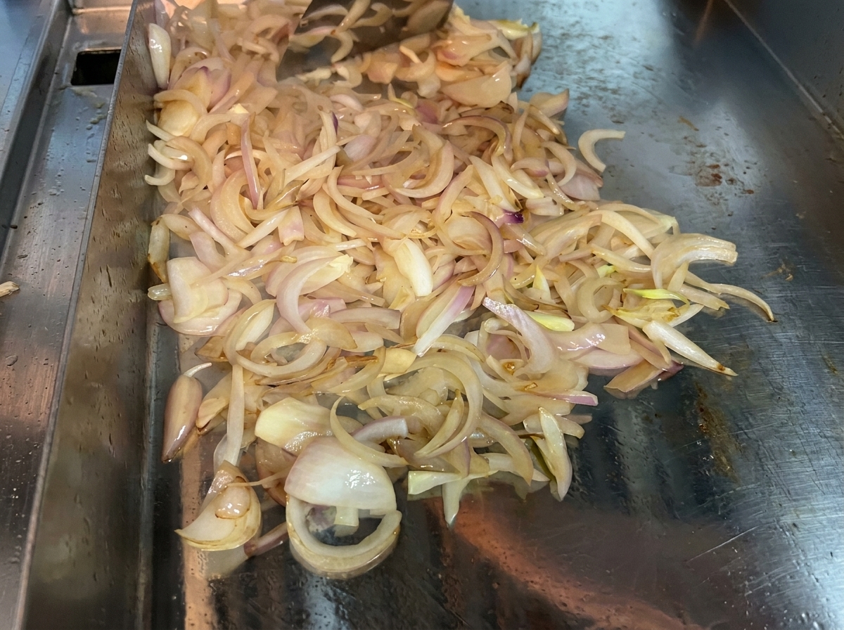 Sliced onions being continuously stirred with a spatula on a hot flat cooking surface to soften and caramelize.