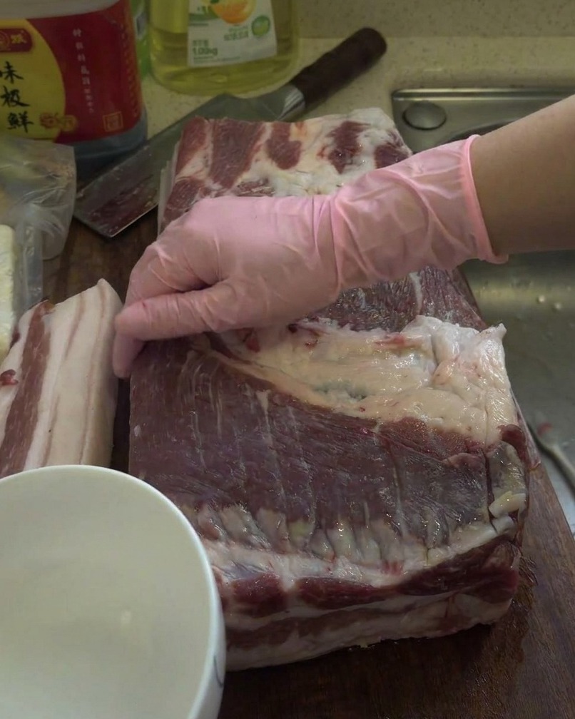 A large raw piece of pork belly being rubbed with white liquor on a wooden cutting board.