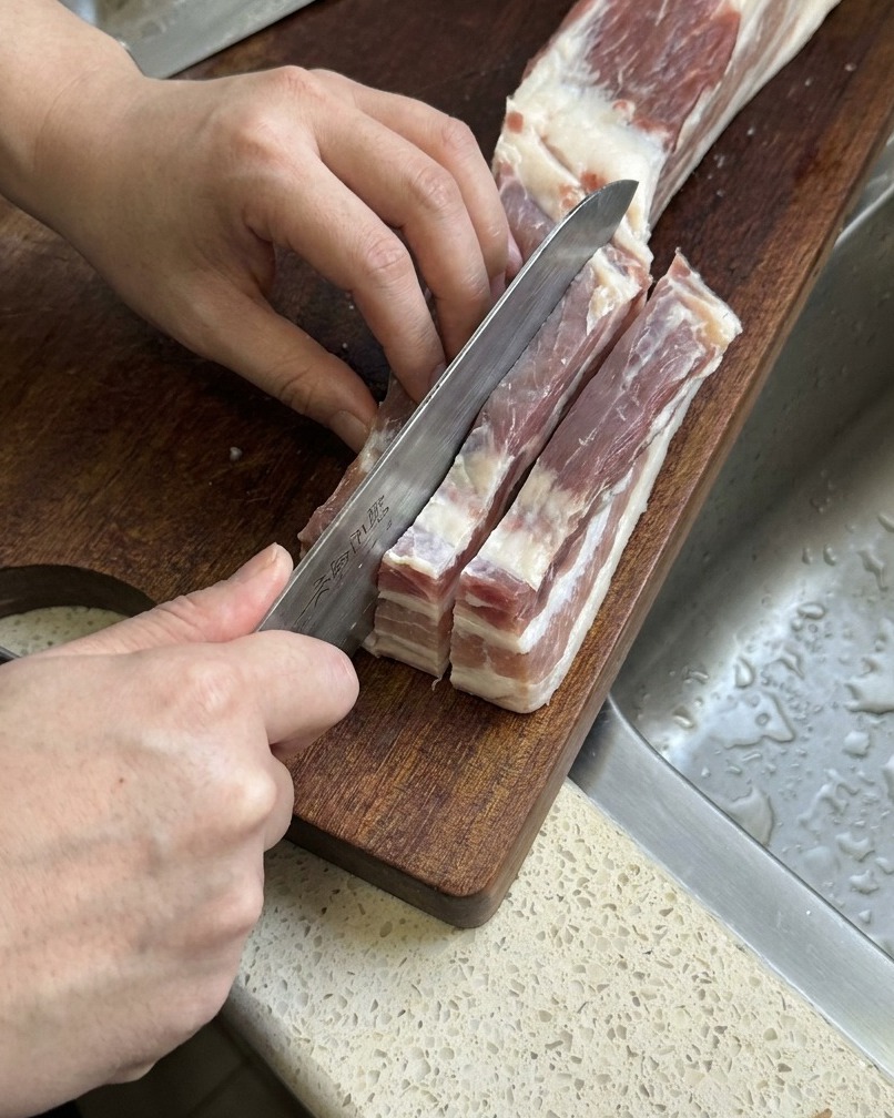 Raw pork belly being sliced into long, thick strips with a sharp kitchen knife.