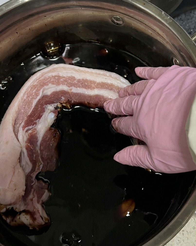 Sliced raw pork belly strips being submerged in a pot of dark soy sauce marinade.