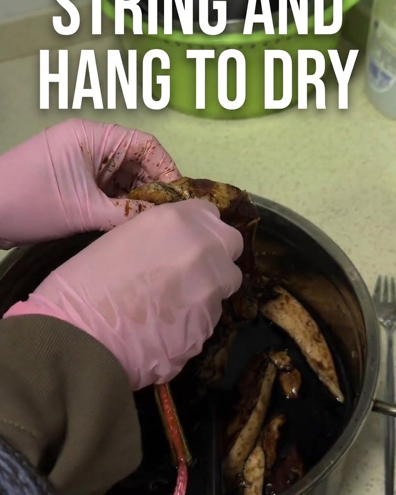 Marinated dark brown pork strip being threaded with a red string for hanging.