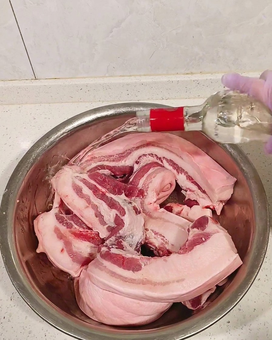 Raw pork belly pieces in a metal bowl being doused with clear white liquor.