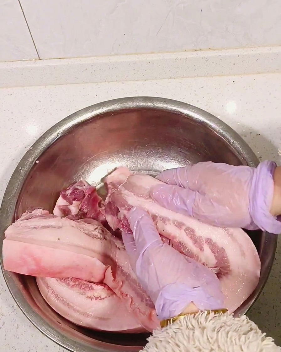 Hands in purple gloves massaging white liquor into raw pork belly.