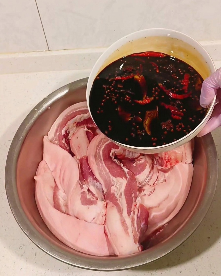 Cooled spice-infused soy sauce being poured over pork belly in a metal bowl.