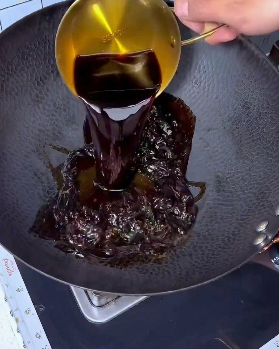 Soy sauce being poured from a golden measuring cup into a large black wok.