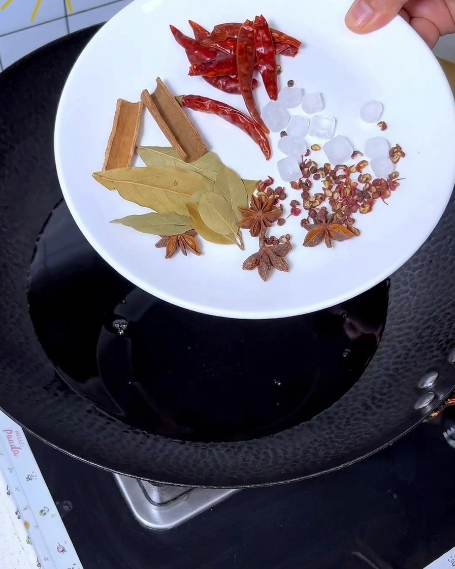 Spices like cinnamon, star anise, bay leaves, chili, and rock sugar being added to the dark liquid in the wok.
