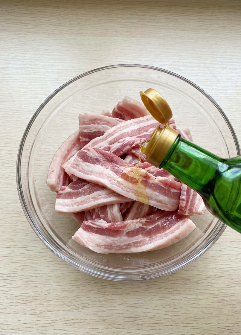 Soy sauce being poured from a glass bottle over raw pork belly strips in a bowl.